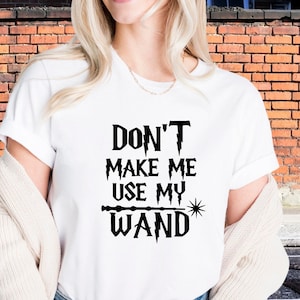 May include: White t-shirt with the text "DON'T MAKE ME USE MY WAND" in black. The design includes a wand and a starburst. The shirt is a classic crew neck style.