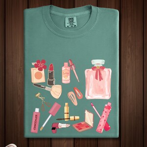 May include: A teal t-shirt with a graphic design of various makeup items, including lipstick, blush, mascara, perfume, and eyeshadow. The design is in pink, red, and gold tones.