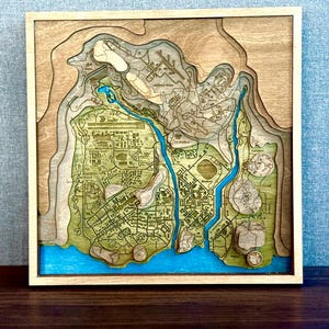 May include: A wooden, layered map with a light brown frame. The map features a detailed, laser-cut design with varying shades of brown, green, and blue. The blue areas represent water, and the green areas represent land. The map is a topographic representation of a city.