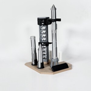 May include: A 3D printed model of a rocket launch site with three rockets on a wooden base. The rockets are silver and black, and the base is a light brown color.