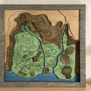 May include: A framed, layered wooden map with a detailed city layout. The map features shades of green, brown, and blue, depicting land and water features. The frame is a muted gray, and the overall aesthetic is a blend of art and cartography.