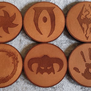 Puede incluir: Seis posavasos redondos y marrones con varios diseños grabados. Los diseños incluyen una luna creciente y una estrella, un símbolo en forma de ojo, un diamante con un dragón, un diseño circular, un casco con cuernos y una huella de mano. Los posavasos son de material marrón claro.