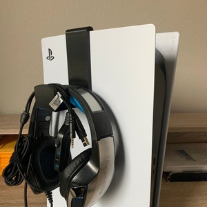 May include: A white PlayStation 5 console with a black headset stand and a black and silver gaming headset. The headset is hanging on the stand, which is attached to the console. The console has the PlayStation logo.
