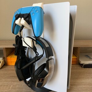 May include: A white gaming console stand with a blue controller and black headphones. The stand has a base for the console and a hook for the controller. The headphones are hanging on the side of the stand.
