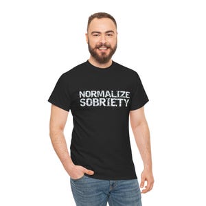 Può includere: T-shirt nero con la scritta "NORMALIZE SOBRIETY" in lettere bianche sbiadite. La maglietta è un classico girocollo. La persona indossa jeans blu.
