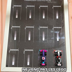 Może przedstawiać: Oprawiona grafika z projektem toru wyścigowego i dziesięcioma ponumerowanymi pasami. Dwa zabawki samochodów wyścigowych, jeden fioletowy i różowy, drugi srebrny i czerwony, znajdują się na dole. Tekst na dole brzmi "NE VEND PAS LES LEGO".