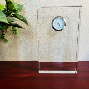 May include: A clear glass clock with a silver face and black hands. The clock is sitting on a wooden surface.