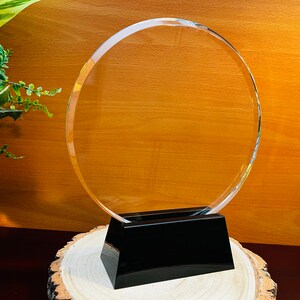May include: A clear glass circle award plaque with a black base. The plaque is on a wooden base.