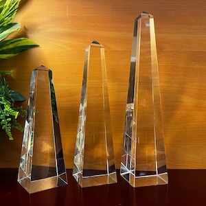 May include: Three clear glass obelisks of varying heights. Each has a pyramidal top and a faceted design, reflecting light. The obelisks are displayed on a dark surface against a warm wood-toned background, with greenery in the corner.