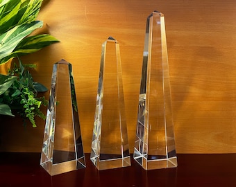 May include: Three clear glass obelisks of varying heights. Each has a pyramidal top and a faceted design, reflecting light. The obelisks are displayed on a dark surface against a warm wood-toned background, with greenery in the corner.
