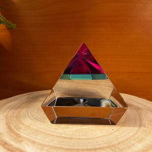 May include: A crystal pyramid with a mirrored finish and a layered design. The top layer is a vibrant pink, the middle layer is a teal green, and the bottom layer is a clear crystal.