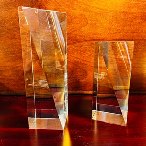 May include: Two clear glass rectangular prisms with beveled edges. The larger prism is on the left and the smaller prism is on the right. Both prisms are sitting on a dark red surface.