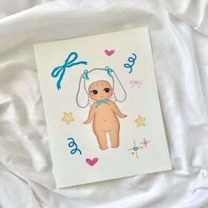 An 8x10 art print of a kawaii baby wearing a white bunny hat with blue bows, surrounded by soft pastel accents. Printed on premium matte paper with a smooth finish and vivid colors. Ships flat in a rigid mailer with protective packaging. Great for kawaii room decor, nurseries, and soft girl aesthetic walls.