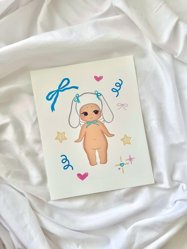 An 8x10 art print of a kawaii baby wearing a white bunny hat with blue bows, surrounded by soft pastel accents. Printed on premium matte paper with a smooth finish and vivid colors. Ships flat in a rigid mailer with protective packaging. Great for kawaii room decor, nurseries, and soft girl aesthetic walls.