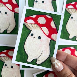 May include: A collection of small, illustrated cards featuring a cartoon mushroom character. The mushroom has a red cap with white spots, a beige stem, and a cute, expressive face. The background is green.