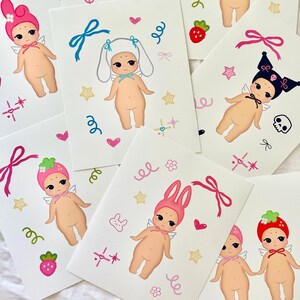 May include: A collection of white cards featuring illustrations of cherubic figures with wings, adorned with various hats and accessories. Each card showcases a unique design, including strawberries, bows, and stars, with a whimsical and playful aesthetic.