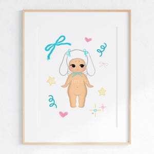 May include: A framed art print featuring a cartoon illustration of a nude figure wearing a white bunny ear hat and a blue bow. The artwork includes blue bows, pink hearts, yellow stars, and blue swirls. The print is in a wooden frame.