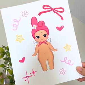May include: A print featuring a cartoon baby angel with a pink hat and bow. The illustration includes pink hearts, stars, flowers, and swirls. The baby angel has small white wings and a bow around its neck.