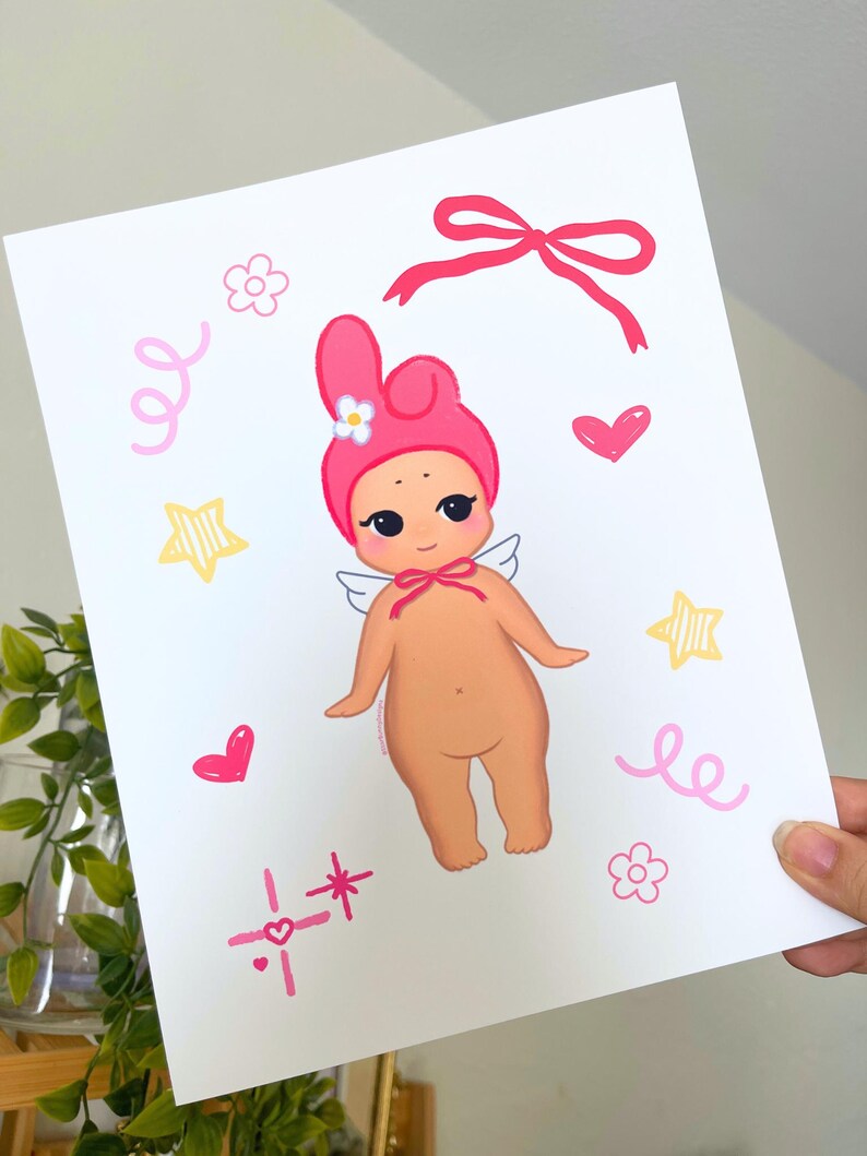May include: A print featuring a cartoon baby angel with a pink hat and bow. The illustration includes pink hearts, stars, flowers, and swirls. The baby angel has small white wings and a bow around its neck.