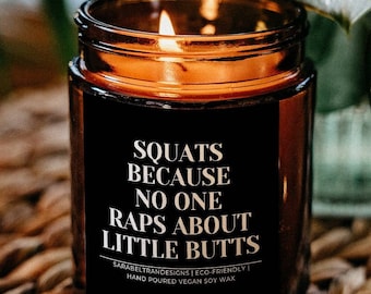 May include: A lit candle in a brown glass jar with the text "Squats Because No One Raps About Little Butts" on a woven basket. The candle is surrounded by green plants and a glass vase.