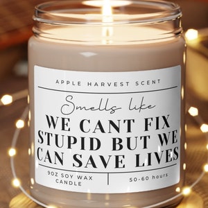 May include: A lit candle in a clear glass jar with a white label that reads "Smells like WE CANT FIX STUPID BUT WE CAN SAVE LIVES." The candle is on a wooden coaster. The candle is an apple harvest scent.