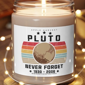 May include: A lit soy wax candle in a glass jar with a label that reads "PLUTO" with a planet graphic and the text "NEVER FORGET 1930-2006". The candle sits on a wooden coaster. The candle is scented with apple harvest.