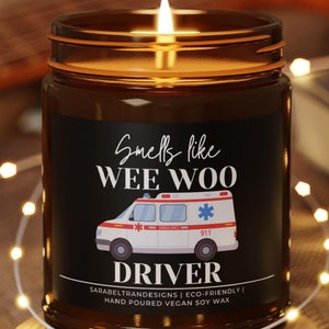May include: A lit amber glass candle with a black label that reads "Smells like WEE WOO" and features an ambulance graphic. The candle sits on a wooden coaster, with string lights and a guitar in the background.