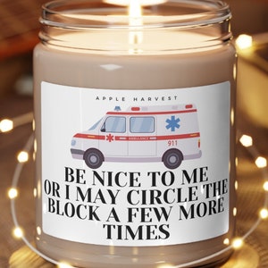 May include: A lit candle in a glass jar with a label that reads "APPLE HARVEST" and features an ambulance graphic. The text on the label says "BE NICE TO ME OR I MAY CIRCLE THE BLOCK A FEW MORE TIMES."