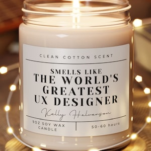 May include: A lit soy wax candle in a glass jar with a label that reads "Smells Like The World's Greatest UX Designer." The candle sits on a wooden coaster, surrounded by string lights. The candle is scented with clean cotton.