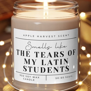 May include: A brown soy wax candle with a white label that reads "Smells like THE TEARS OF MY LATIN STUDENTS" and "APPLE HARVEST SCENT". The candle is on a wooden coaster and surrounded by string lights.
