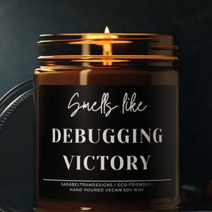 May include: A lit candle in a brown glass jar with a black label. The label reads "Smells like Debugging Victory". The jar has a silver lid and is placed on a dark surface. The candle is hand poured with vegan soy wax.