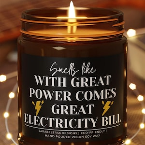 May include: A brown glass candle with a black label that reads "Smells like WITH GREAT POWER COMES GREAT ELECTRICITY BILL". The candle is lit and sitting on a wooden coaster.