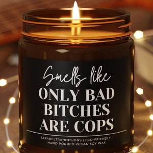 May include: A lit candle in a brown glass jar with a black label. The label reads "Smells like ONLY BAD BITCHES ARE COPS." The candle sits on a small wooden coaster, with string lights and a guitar in the background.