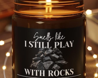 May include: A lit soy wax candle in a brown glass jar with a black label that reads "Smells like I STILL PLAY WITH ROCKS". The candle sits on a wooden coaster, with string lights and a guitar in the background.