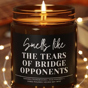 May include: A lit soy wax candle in a brown glass jar with a black label that reads "Smells like the Tears of Bridge Opponents." The candle sits on a wooden coaster, with fairy lights and a guitar in the background.