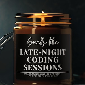Puede incluir: Una vela encendida en un frasco de vidrio marrón con una etiqueta negra. La etiqueta dice "Smells like LATE-NIGHT CODING SESSIONS". El frasco tiene una tapa de metal y está sobre una superficie oscura. Cera de soja vegana vertida a mano.