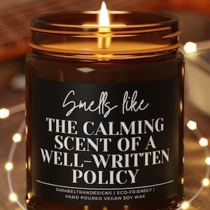 May include: A lit brown glass jar candle on a wooden coaster. The candle has a black label with white text that reads "Smells like the calming scent of a well-written policy." The candle is surrounded by fairy lights.