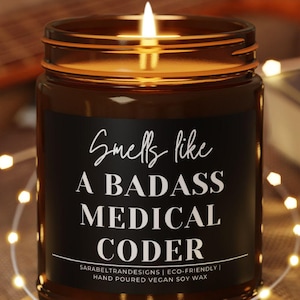 May include: A lit brown glass candle with the text "Smells like A BADASS MEDICAL CODER" on a black label. The candle sits on a small wooden coaster, surrounded by string lights. The background includes a guitar and a book.