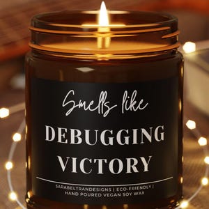 May include: A lit brown glass candle with the text "Smells like Debugging Victory" on a black label. The candle sits on a small wooden coaster, with a string of fairy lights and a guitar in the background.