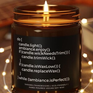 May include: A lit brown glass candle with a black label featuring code-like text. The candle sits on a wooden coaster, surrounded by fairy lights. The text on the label reads "SARABELTRANDESIGNS | ECO-FRIENDLY | HAND POURED VEGAN SOY WAX".
