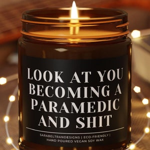 May include: A lit candle in a brown glass jar with a black label featuring the white text: LOOK AT YOU BECOMING A PARAMEDIC AND SHIT. Below this, smaller text reads: SARABELTRANDESIGNS | ECO-FRIENDLY | HAND POURED VEGAN SOY WAX. The jar sits on a wooden coaster.