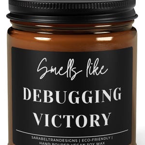 May include: A brown glass jar candle with a black lid. The candle has a black label with white text that reads "Smells like Debugging Victory". The bottom of the label says "SARABELTRANDESIGNS | ECO-FRIENDLY | HAND POURED VEGAN SOY WAX".