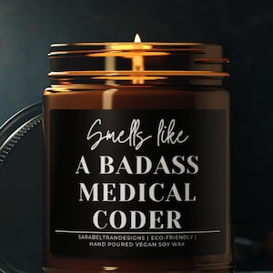 May include: A lit candle in a brown glass jar with a black label. The label reads "Smells like A BADASS MEDICAL CODER". The jar has a silver lid next to it. The candle is hand-poured vegan soy wax.