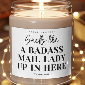 May include: A lit candle in a clear glass jar with a white label that reads "Smells like A BADASS MAIL LADY UP IN HERE". The candle sits on a small wooden coaster. The candle is surrounded by string lights.