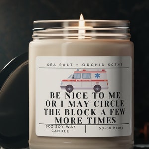 May include: A beige soy wax candle with a white label that reads "SEA SALT ORCHID SCENT" and features a cartoon ambulance with the text "BE NICE TO ME OR I MAY CIRCLE THE BLOCK A FEW MORE TIMES". The candle is 9 oz and has a burn time of 50-60 hours.