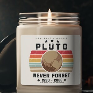 May include: A lit candle in a glass jar with a label that reads "PLUTO NEVER FORGET 1930-2006". The label has a retro design with a planet illustration and the words "Sea Salt & Orchid". The candle is on a dark surface.