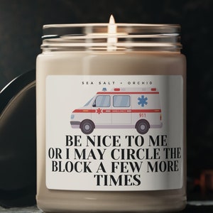 May include: A beige candle with a white label that says "SEA SALT ORCHID" and features a cartoon ambulance with the text "BE NICE TO ME OR I MAY CIRCLE THE BLOCK A FEW MORE TIMES".