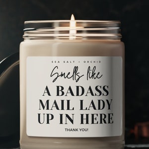May include: A lit, beige-colored candle in a glass jar with a black lid. The label reads "Smells like A BADASS MAIL LADY UP IN HERE THANK YOU!" The candle is labeled "SEA SALT + ORCHID".