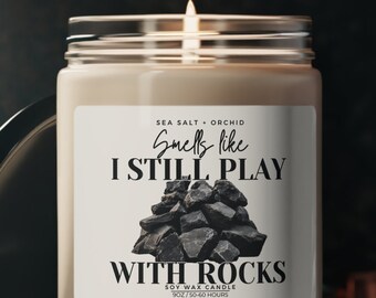 May include: A lit soy wax candle in a clear glass jar with a black lid. The label reads "Smells like I Still Play With Rocks." The candle has a white label with black text and a graphic of rocks. The scent is sea salt and orchid.