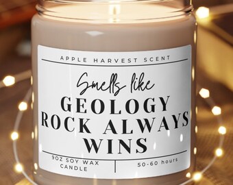 May include: A 9oz soy wax candle in a clear glass jar with a white label that reads Apple Harvest Scent, Smells like GEOLOGY ROCK ALWAYS WINS, and 50-60 hours. The candle sits on a round wooden coaster and is lit with a visible flame.